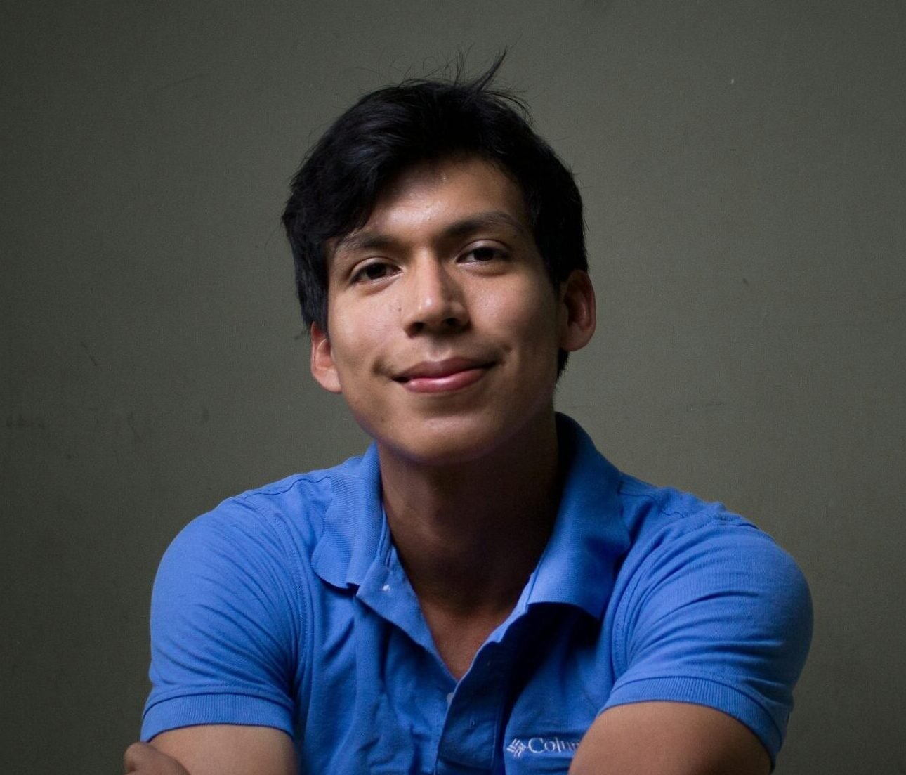 Young Hispanic man smiling confidently in a blue shirt against a dark background.